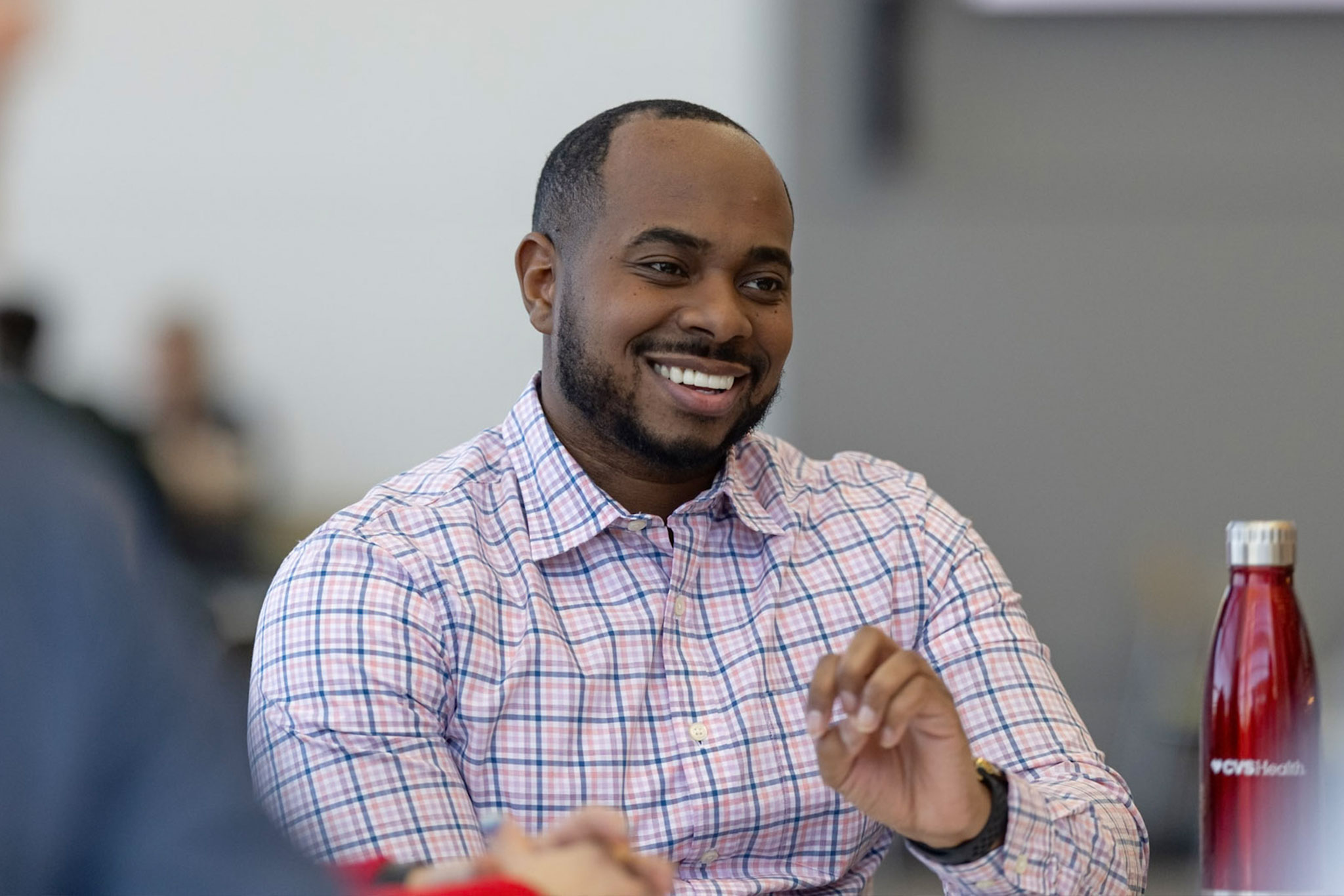 A man in a checked shirt smiles while sitting at a table in a meeting or informal discussion. There is a red stainless steel bottle labeled CVS Health on the table. The background is blurred, focusing attention on the man's pleasant expression.