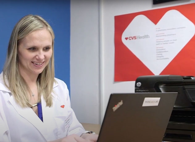 A woman in a white lab coat is seated and smiling while looking at a laptop. The laptop has a CVS Health sticker on it. Behind her, a wall poster featuring a heart shape and the CVS Health logo is visible. The environment suggests a professional or medical setting.