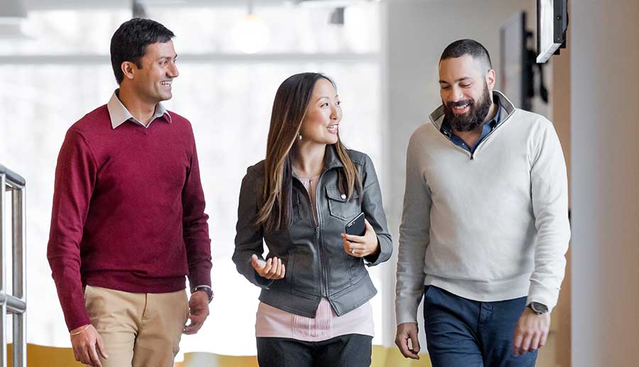 Three people are walking together in a well-lit indoor setting. They appear to be engaged in a conversation and are smiling. The person on the left is wearing a red sweater, the person in the middle has a gray jacket, and the person on the right is in a white sweater.