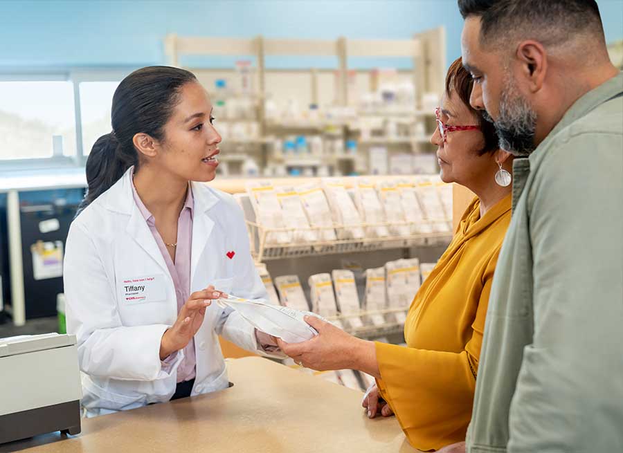 A pharmacist, wearing a white lab coat and name tag, is engaged in conversation with a female customer holding a package. A male customer stands beside the woman, listening attentively. They are in a pharmacy with shelves of medications in the background.