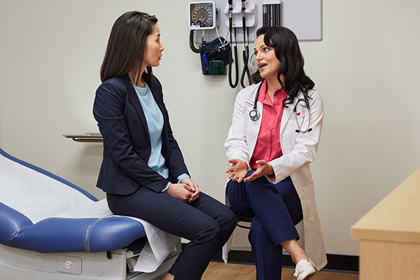 A doctor in a white coat talks with a woman sitting on an exam table in a medical office. Medical equipment hangs on the wall behind them, and they appear engaged in conversation.