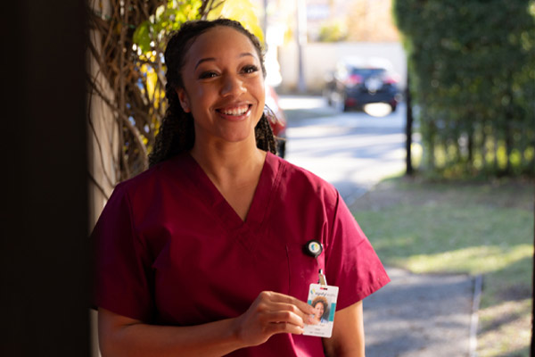 A smiling woman in maroon scrubs stands outside, holding up her ID badge. Sunlight and greenery are visible in the background, suggesting a residential neighborhood.
