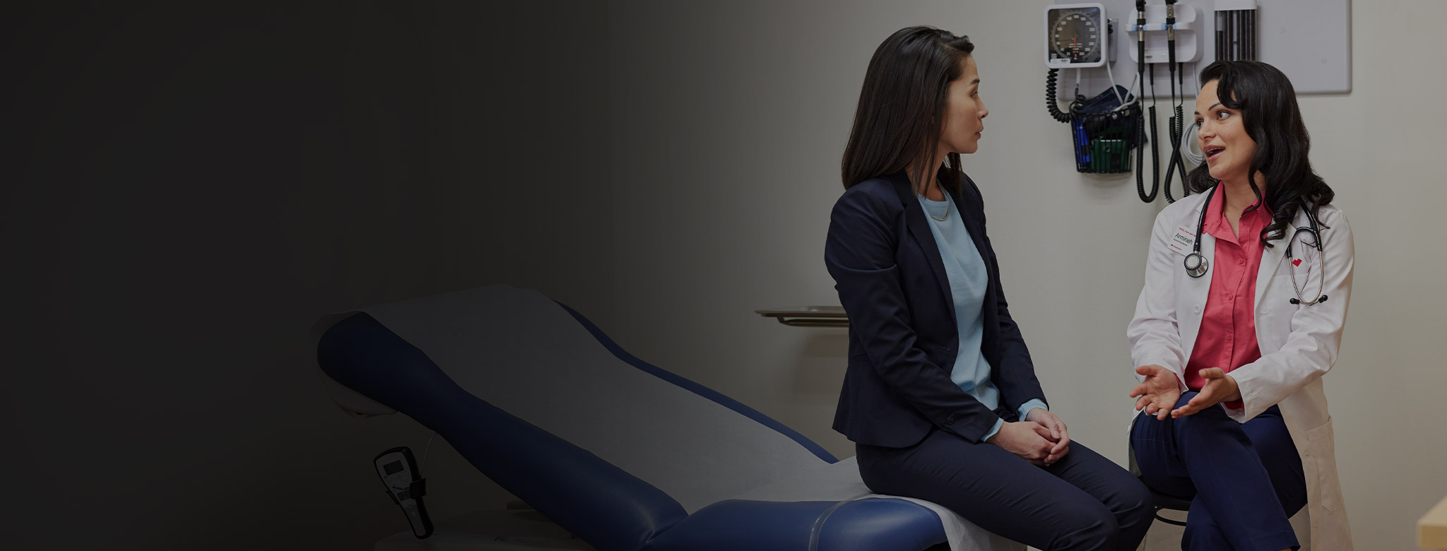 A doctor sits and talks with a patient in a medical exam room. The doctor gestures with her hands while the patient listens attentively. Medical equipment hangs on the wall in the background.