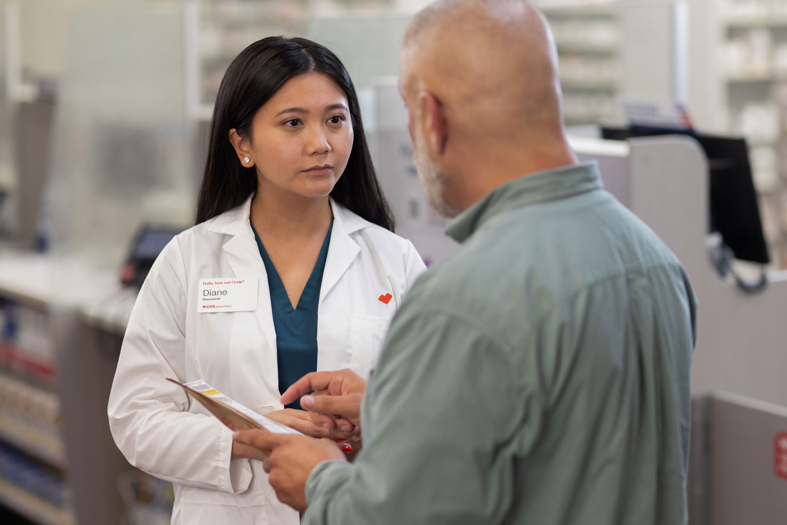 A female pharmacist wearing a white lab coat and name tag listens attentively to a male customer who is holding a prescription bag. They are standing in a pharmacy, with shelves of medication visible in the background.