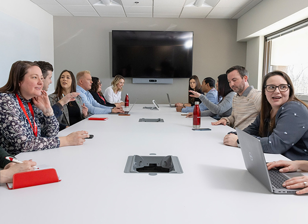 A diverse group of people is seated around a long, white conference table in a modern office, engaging in discussion. Some have laptops and notebooks, while others gesticulate or listen. A large screen is mounted on the wall at the far end of the room.