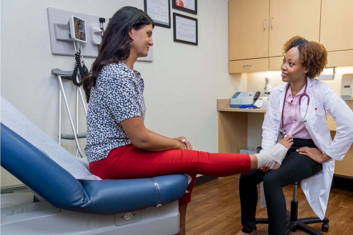 A doctor wearing a white coat is kneeling and examining a patient's bandaged foot in a medical clinic. The patient is sitting on an examination table, wearing a patterned shirt and red pants. Medical equipment and framed certificates are visible in the background.