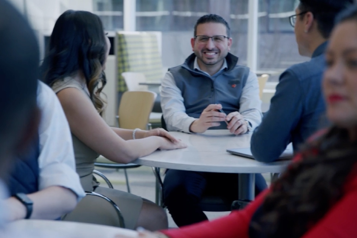 A group of people are gathered around a table in a modern office setting. One person is in focus, smiling and holding a smartphone, while the others are conversing and partially visible. The background features large windows and contemporary furniture.