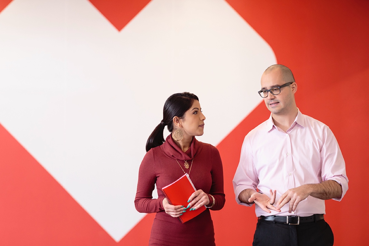 Two people are standing and talking in front of a large red heart backdrop. The woman on the left is holding a red notebook and wearing a red sweater, while the man on the right is wearing glasses and a white dress shirt, gesturing with his hands.