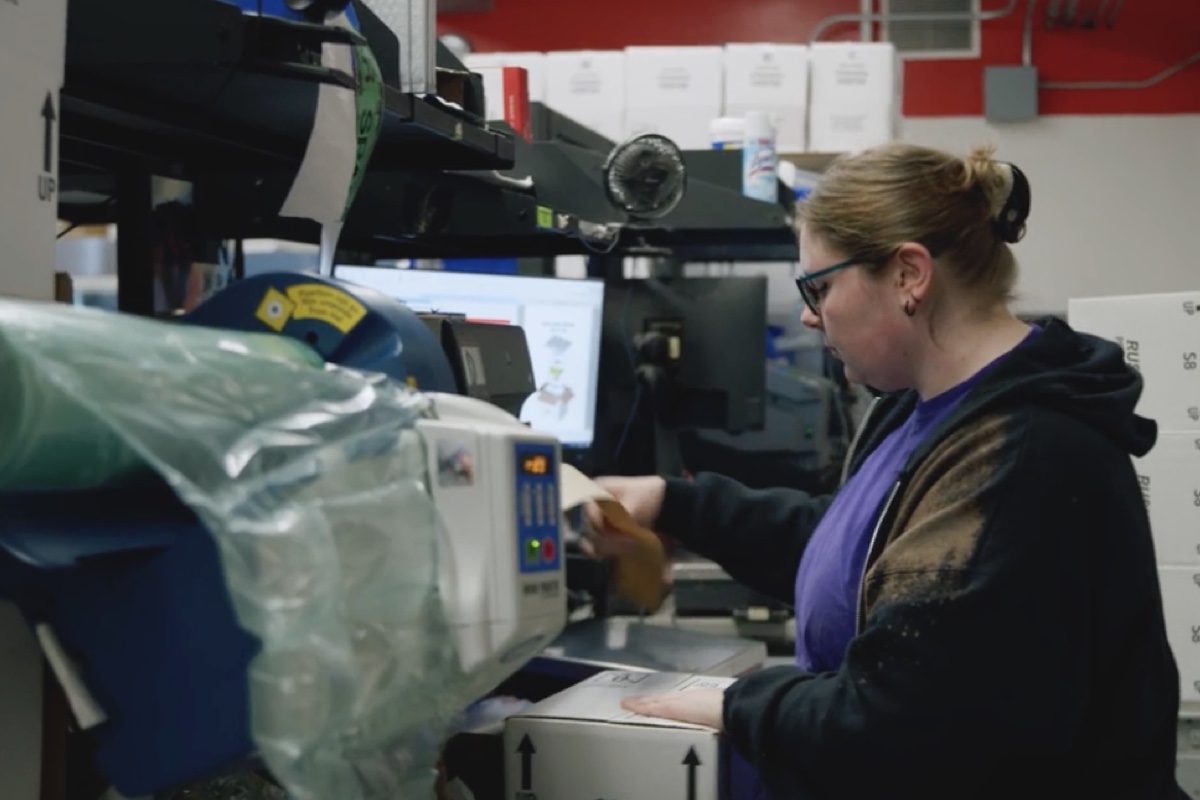 A woman wearing glasses and a black hoodie works with a packaging machine in an industrial setting. She is placing items into a box. The background includes shelves with various supplies and equipment.