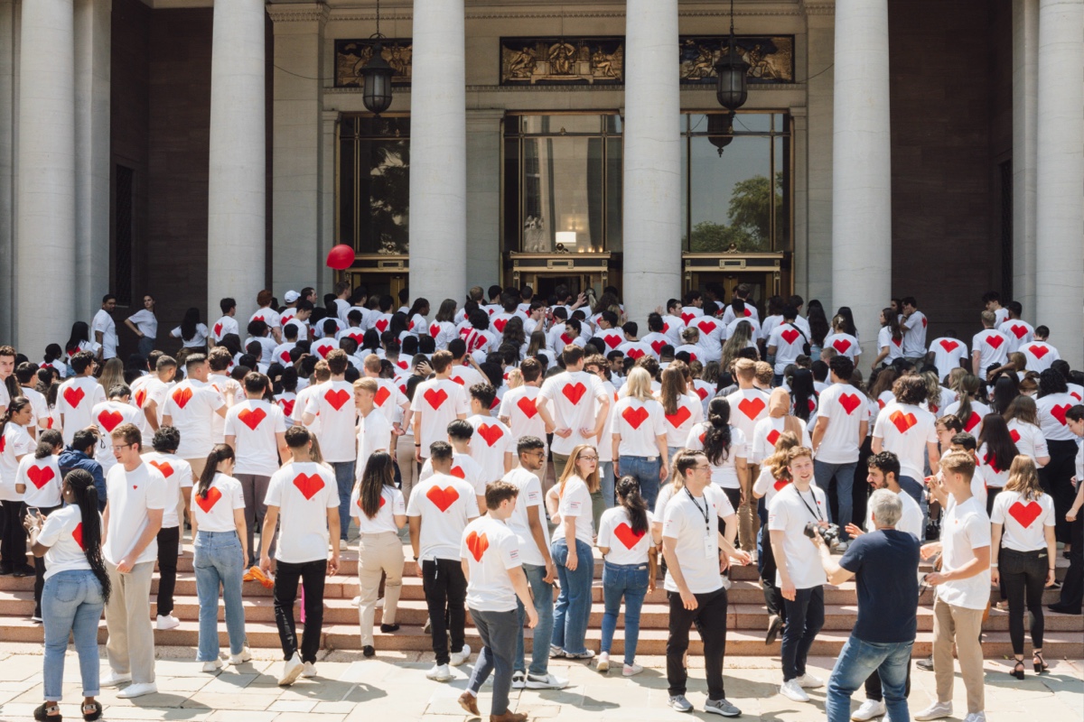 A large group of people wearing white T-shirts with red hearts on the back gather on the steps of a building with columns. Some individuals are holding cameras, and one person is carrying a red balloon. The scene appears to be a social or community event.