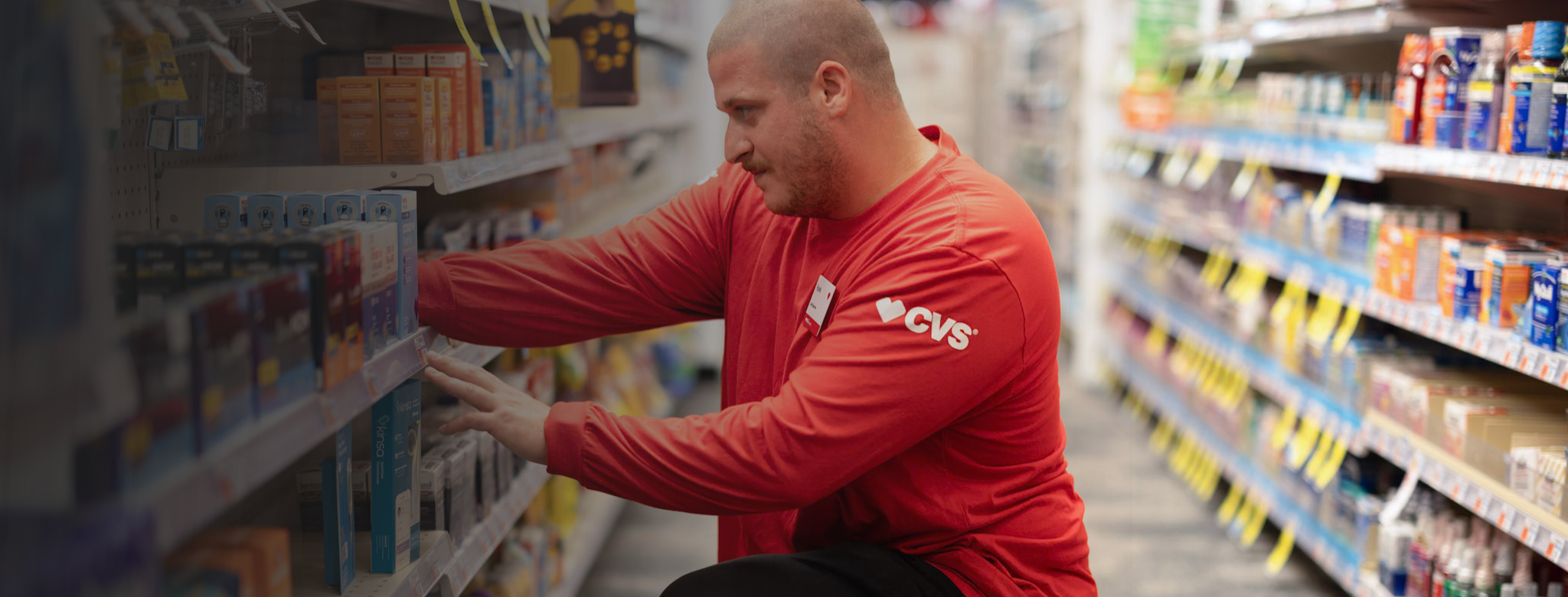 A person wearing a red long‑sleeve shirt with a CVS logo kneels in a retail aisle, restocking or arranging boxed products on a store shelf. Aisles on both sides are filled with health and personal care items, with small yellow price tags visible along the shelves and the background softly out of focus.