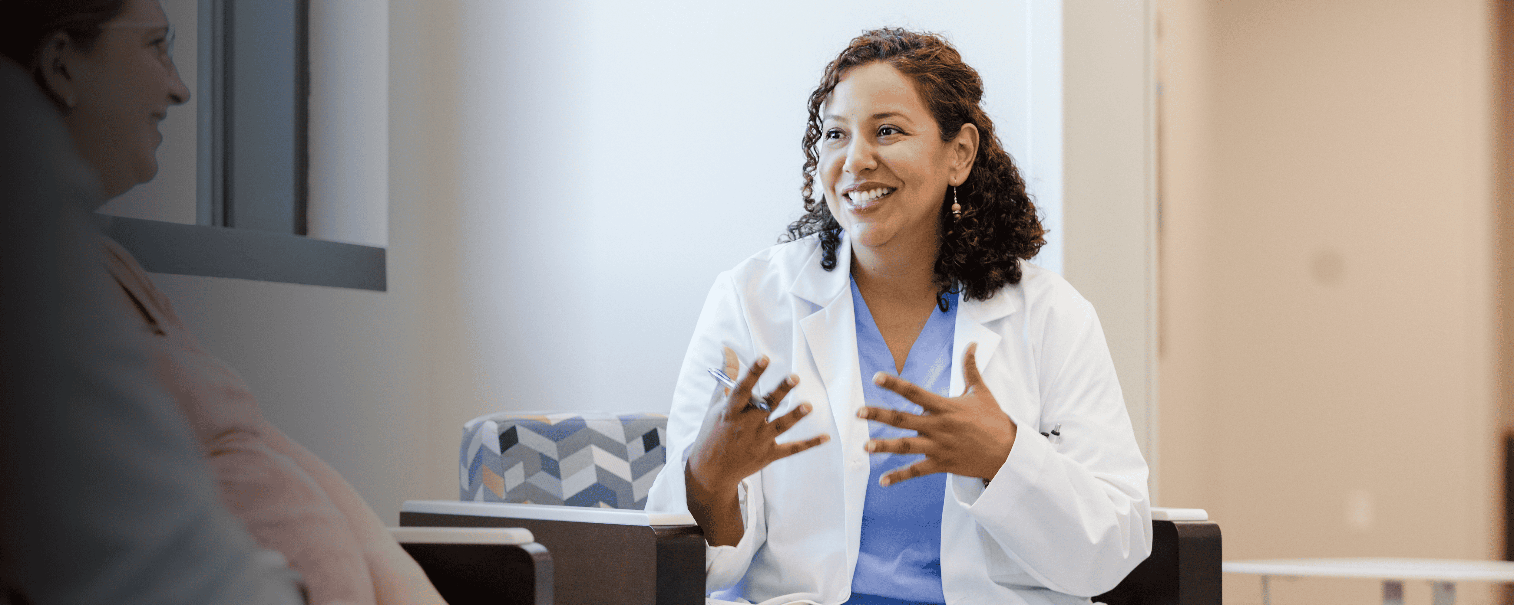 A smiling female healthcare provider in a white coat and blue scrubs is speaking with a patient in a warmly lit clinic setting. She is using expressive hand gestures while seated across from the patient, creating a welcoming and engaging atmosphere.