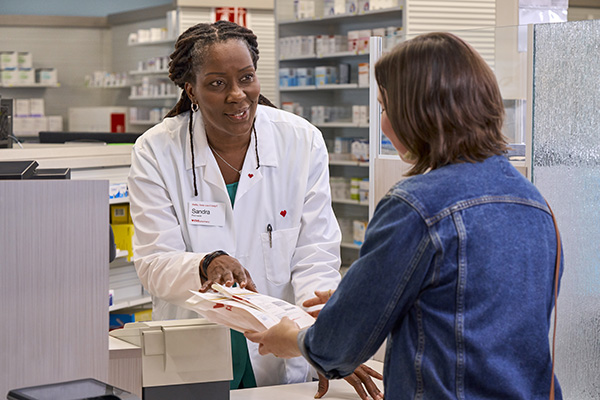 A pharmacist hands a bag of prescriptions to a customer at a pharmacy counter. The pharmacist, wearing a white coat and a name tag, stands behind the counter while the customer, in a denim jacket, reaches out to receive the items.