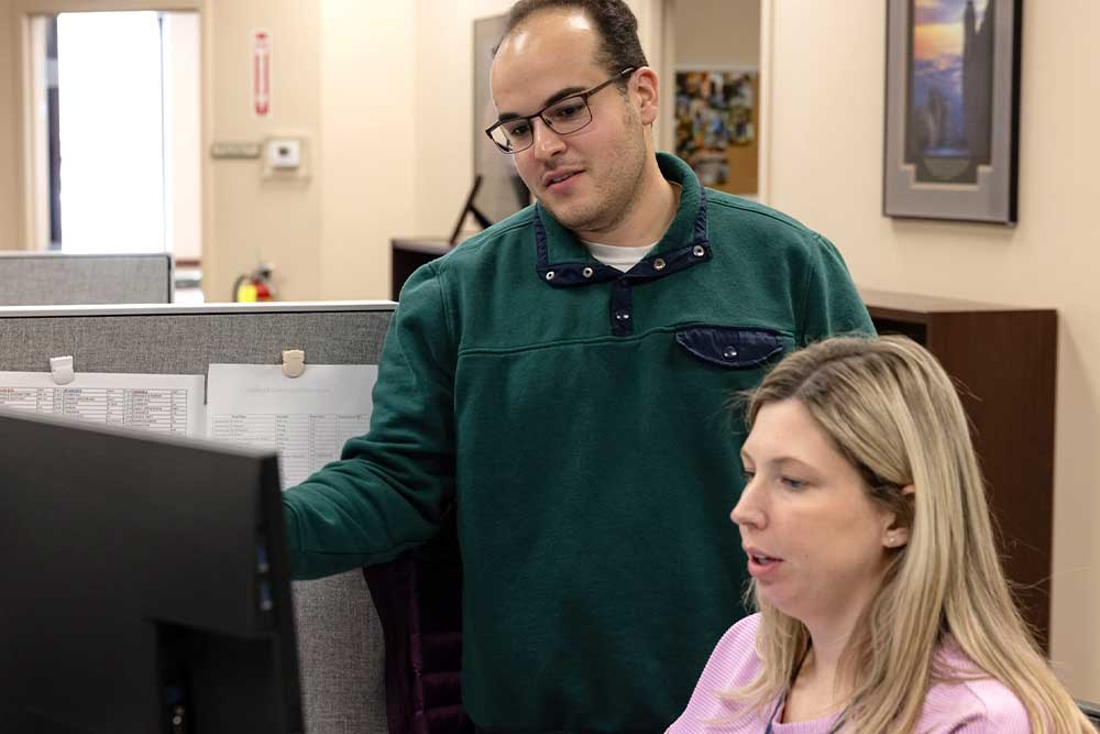 A pharmacist, wearing a white lab coat and a name tag, smiles while handing a prescription medication to a customer. Behind her, shelves are stocked with various medications and pharmaceutical supplies.