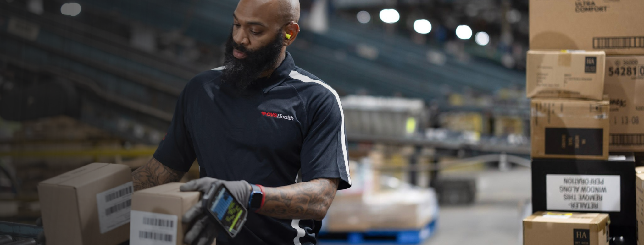 CVS Health distribution center employee scans and sorts shipping boxes on a warehouse floor, with stacked packages, shelving and logistics operations visible in the background.