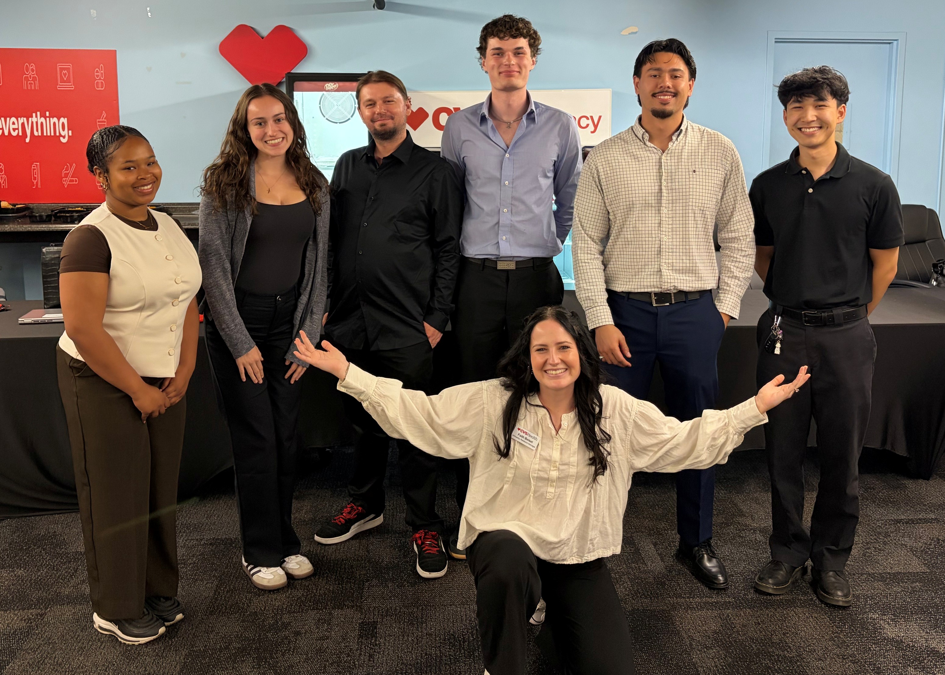 Group photo of seven people in a CVS Pharmacy-branded meeting space. Six people stand in a row while one person kneels in front with arms extended. The group is dressed in business-casual attire and poses in front of a wall featuring CVS Pharmacy signage and a large red heart logo. Tables with black tablecloths are visible in the background, and the room has light blue walls and office-style furnishings.