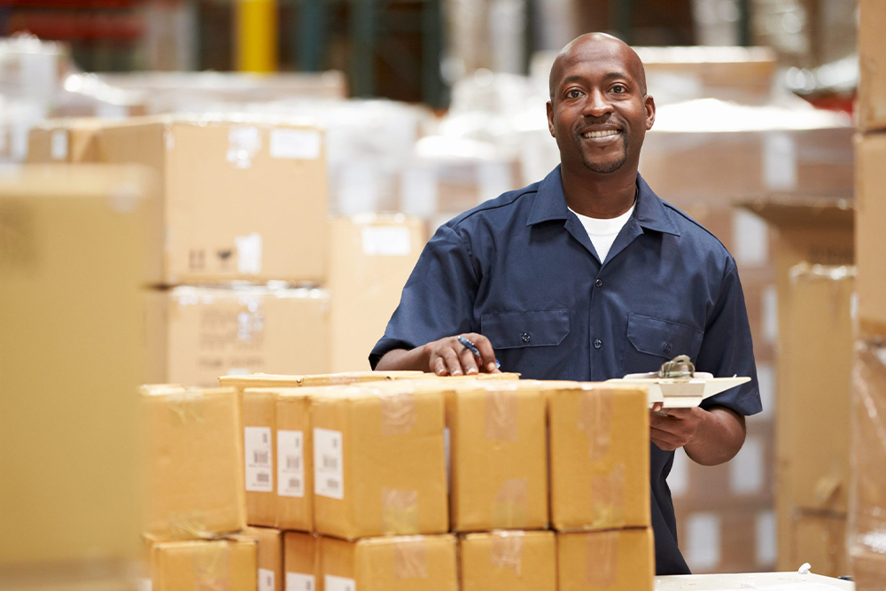 A man in a warehouse standing among stacked cardboard boxes. He is wearing a navy blue work shirt and holding a clipboard with a pen in his right hand. He has a friendly expression, and the background shows more shelves and boxes.