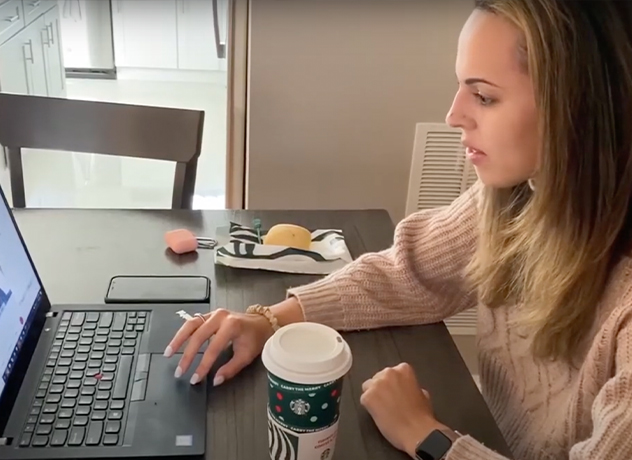 A woman sits at a table, working on a laptop. She has long hair and wears a light-colored sweater. In front of her is a cup of coffee from Starbucks. Beside the laptop, there is a pair of glasses and a small object resembling a snack on the table.