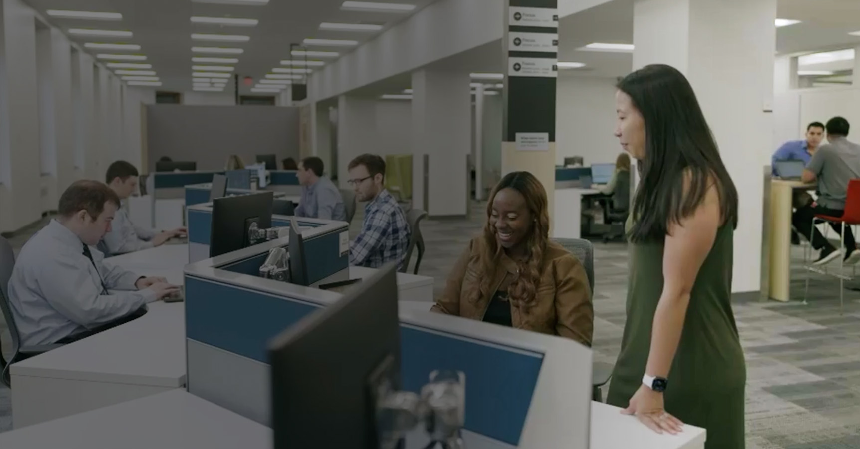 A bustling office scene with workers sitting at desks, typing on computers. A woman with long dark hair is standing and talking to another woman with long hair seated at a desk. The office layout is open with rows of cubicles and bright overhead lighting.