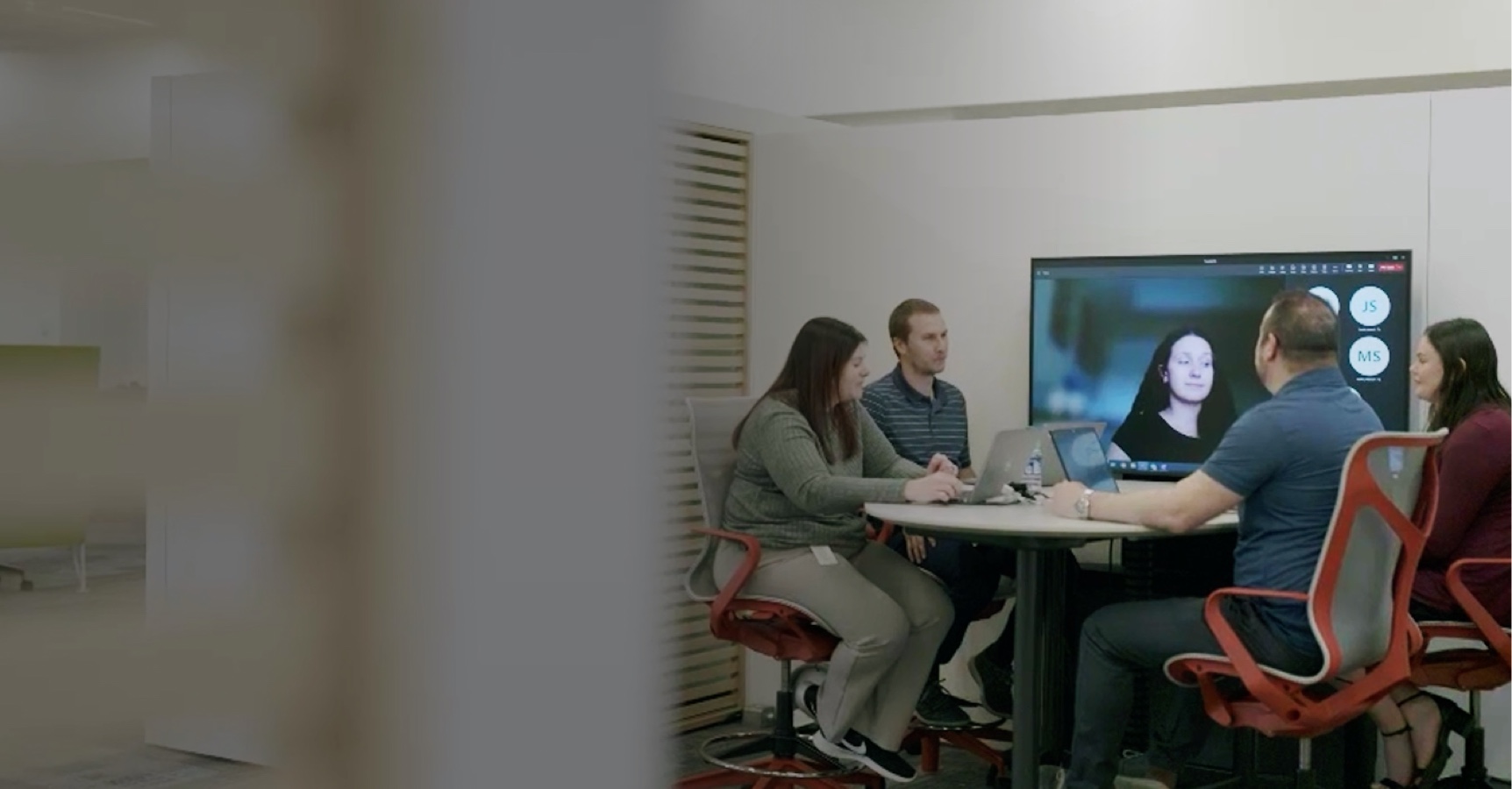 Four people are sitting around a conference table having a meeting, with one person participating virtually via a video call displayed on a large monitor. The group appears engaged in discussion, seated in an open and modern office space.