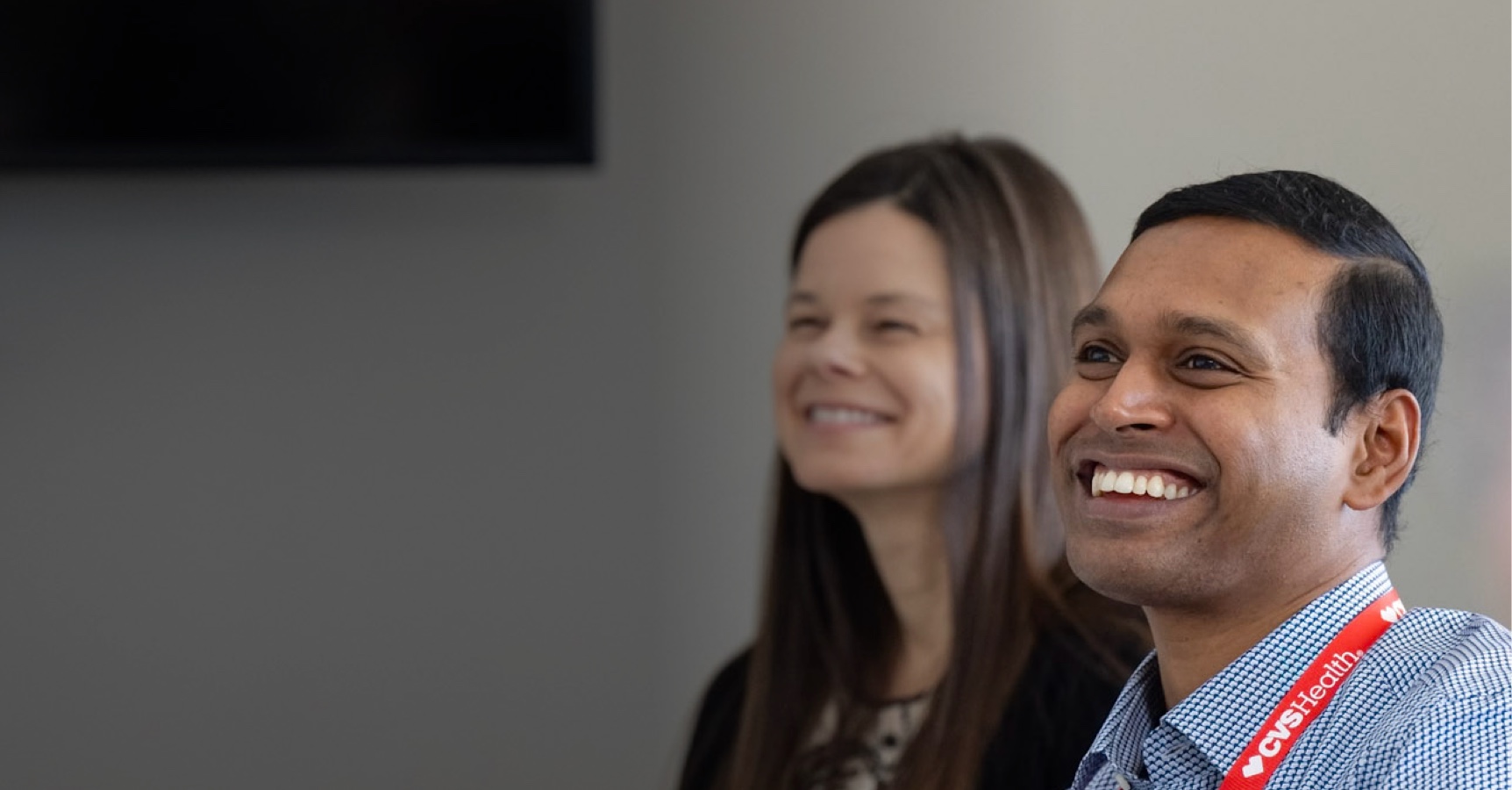 Two people are smiling. The person in the foreground on the right is wearing a red lanyard with "CVS Health" on it. They are both looking towards the left of the frame. The background is plain with a dark object partially visible on the top left.