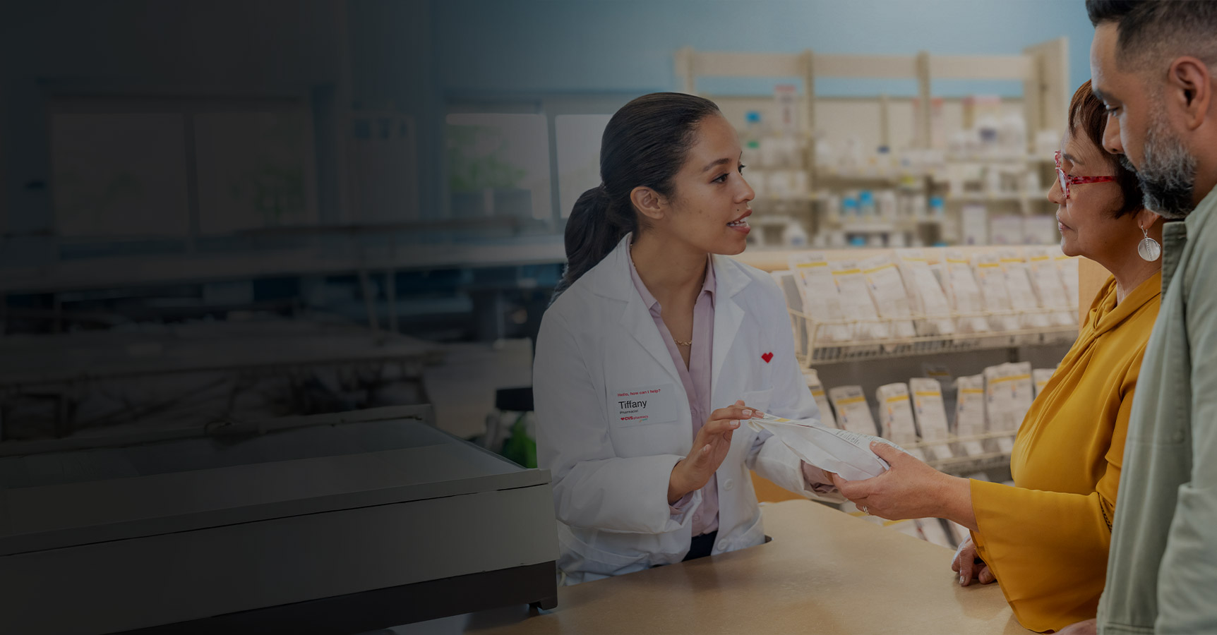 A pharmacist in a white coat speaks to two customers at a counter, handing them a prescription bag in a well-lit pharmacy. Shelves filled with medication are visible in the background.