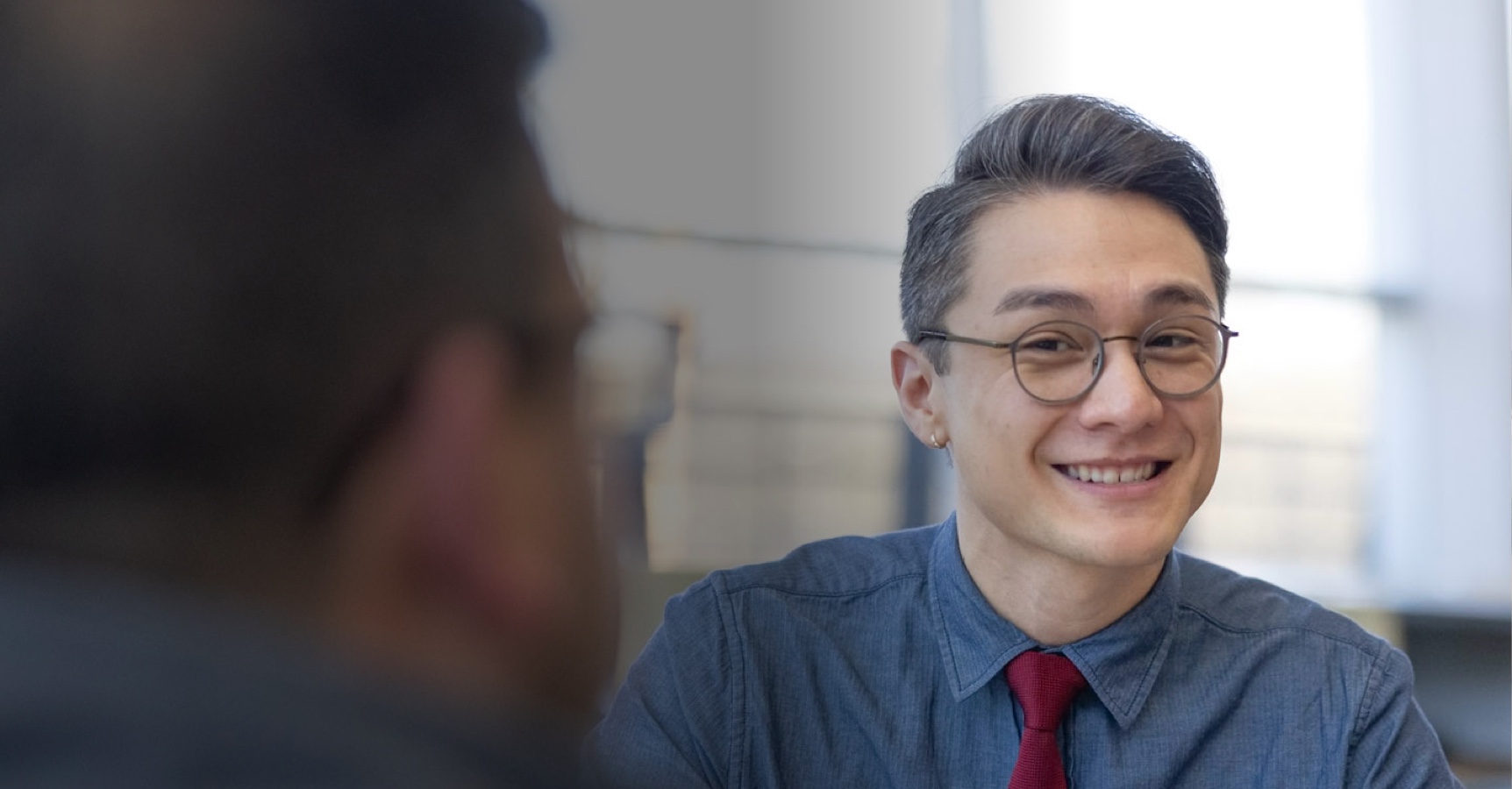 A person with short hair, wearing glasses, a blue collared shirt, and a red tie is smiling in the foreground. Another person's blurred profile is in the foreground. The background is a bright, indoor setting.