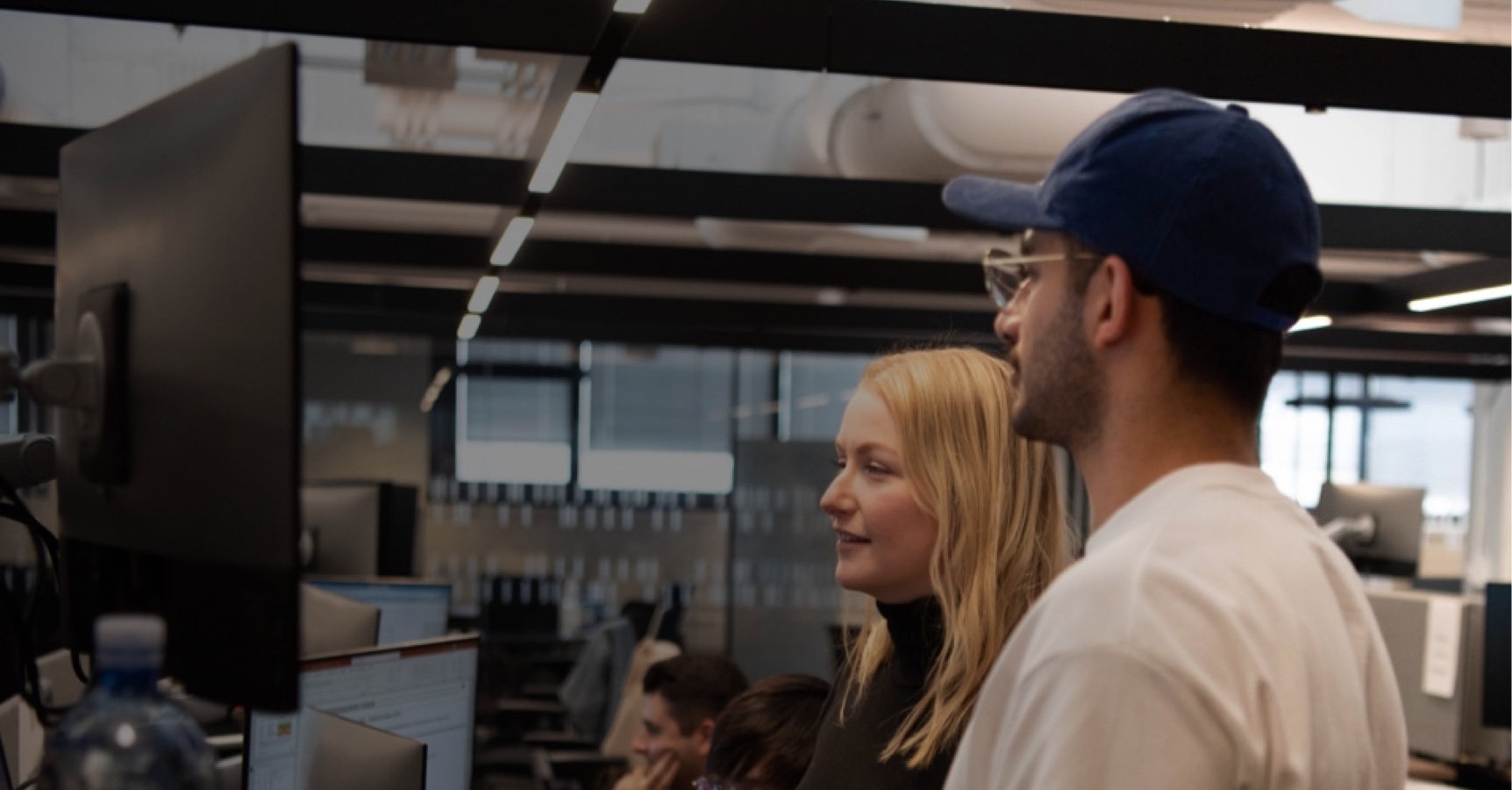 Two people are standing in front of a large computer monitor in an office setting. The person on the left, with blonde hair, and the person on the right, wearing a blue cap and glasses, seem engaged in a discussion or collaboration. Several other people are working in the background.