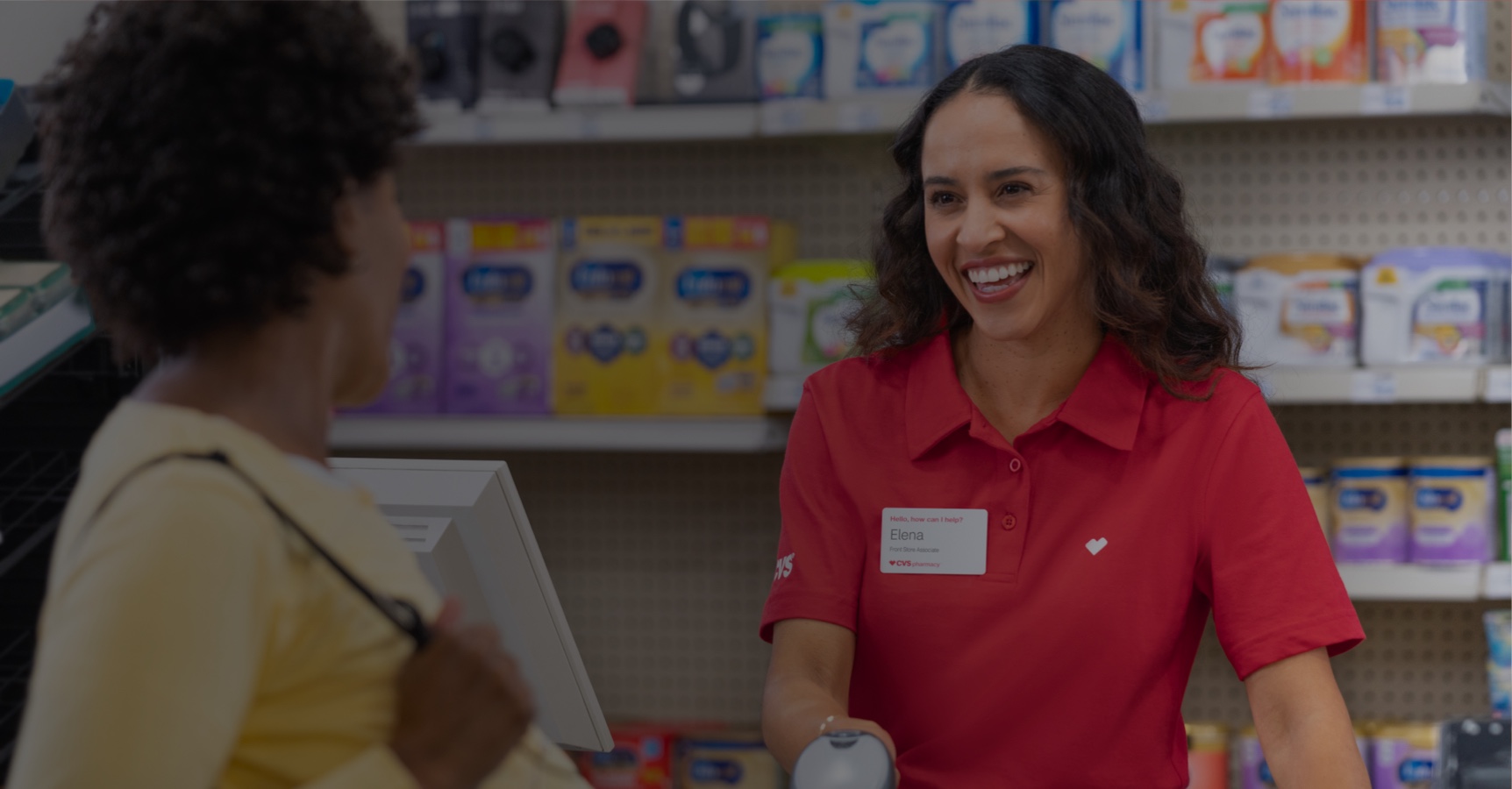 A cashier wearing a red shirt and name tag smiles while scanning items for a customer at a store counter. The customer, holding a shopping basket, is looking at the cashier. Shelves stocked with various products are visible in the background.