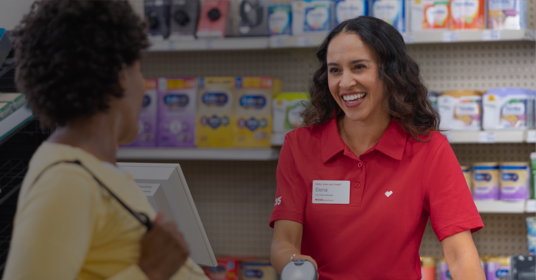 A smiling cashier in a red uniform stands behind a counter, assisting a customer. Shelves in the background display various products. The cashier holds a scanning device and wears a name tag that reads "Elena." The customer, partially visible, holds a bag.