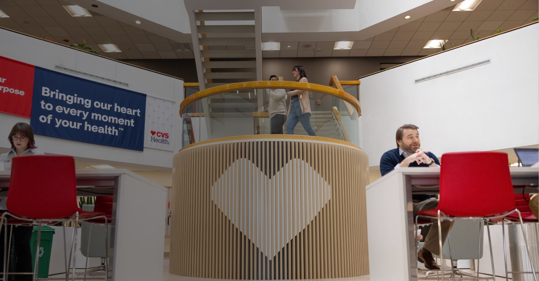 A modern indoor space features a circular wooden structure with a heart design in the center. Two people talk on a staircase above, while individuals sit at tables with red chairs on either side. Banners with 