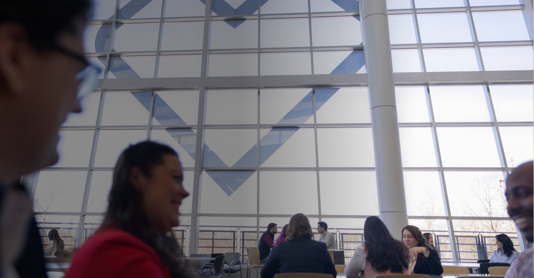 A spacious, modern cafeteria with large windows revealing a heart design on the structure outside. People sit at tables, chatting and eating, with natural light filling the area. The focus is on the interactions and relaxed atmosphere.