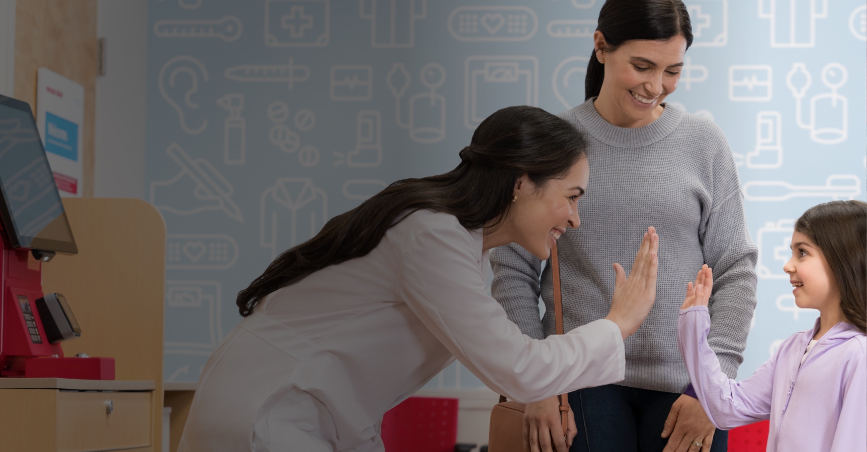 A female pharmacist in a white coat is smiling and giving a high-five to a young girl. The girl's mother, wearing a gray sweater, stands next to her, smiling. They are in a pharmacy with a healthcare-themed wallpaper in the background.