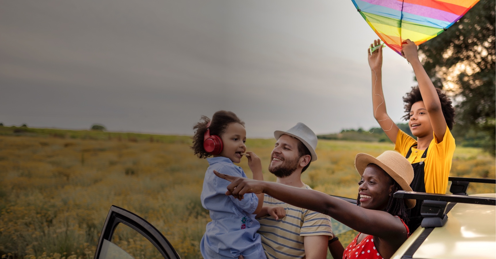 A joyful family enjoys a day outdoors. A woman in a straw hat is standing by an open car door, pointing towards the sky, while a man holds a young girl wearing red headphones. Another child in a yellow shirt is holding up a colorful kite. A field stretches behind them.