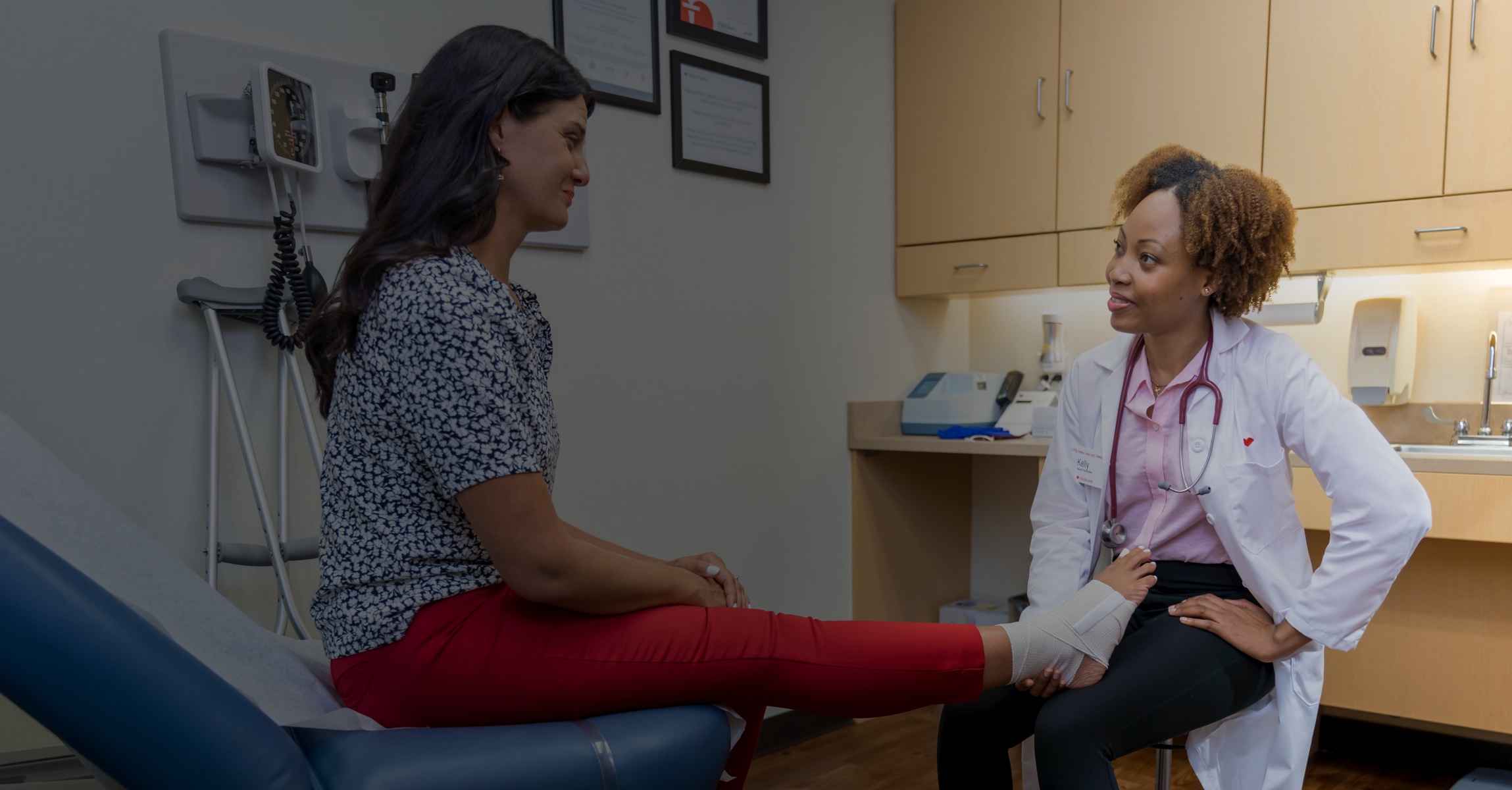 A doctor in a white coat is examining a patient's right foot, which is wrapped in a medical bandage. The patient is seated on an exam table wearing a patterned blouse and red pants. They are in a medical examination room furnished with cabinets and medical equipment.