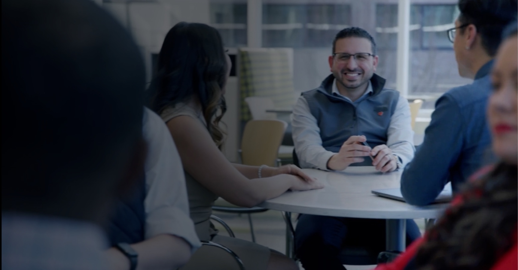 A group of people sit around a white table, engaged in conversation in a modern office setting. One man, wearing glasses and a gray vest, smiles and looks directly ahead while holding a cup. Another person has a laptop open on the table.