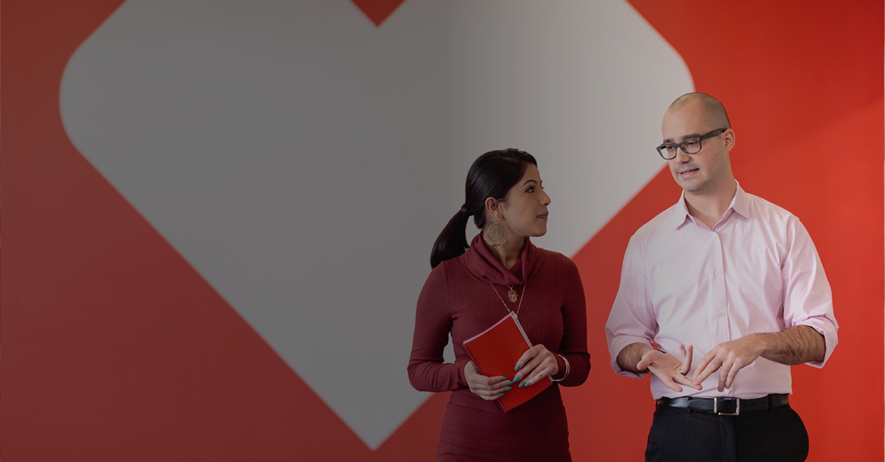 A woman in a red dress holding a notebook and a man in a light pink shirt having a conversation in front of a large red and white heart design on a wall. The woman is looking at the man, and the man appears to be explaining something.