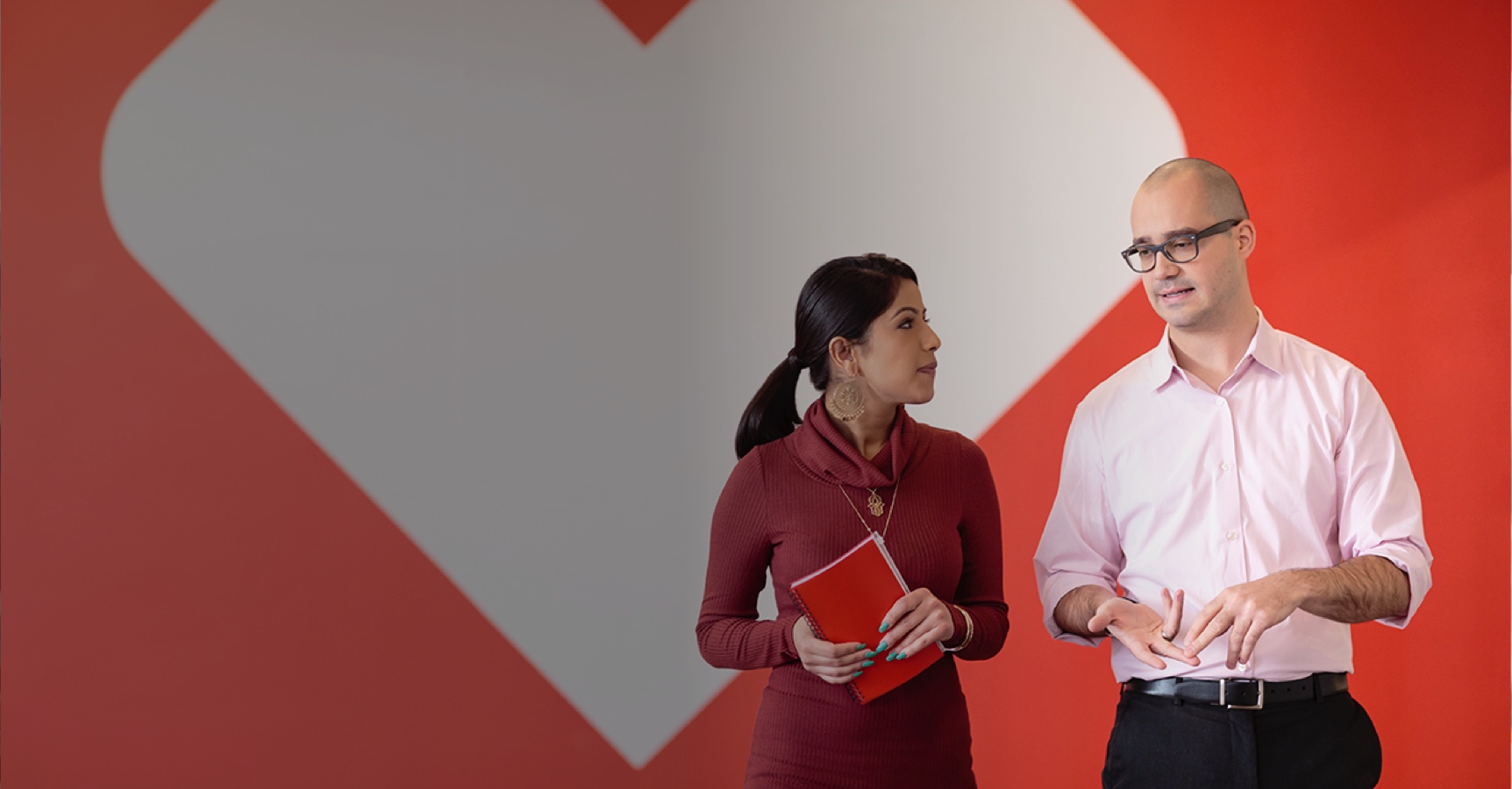 A woman in a red dress holding a red notebook converses with a man in a pale pink shirt and black trousers. They are standing in front of a large red and white heart-shaped backdrop. The man appears to be explaining something while the woman listens attentively.