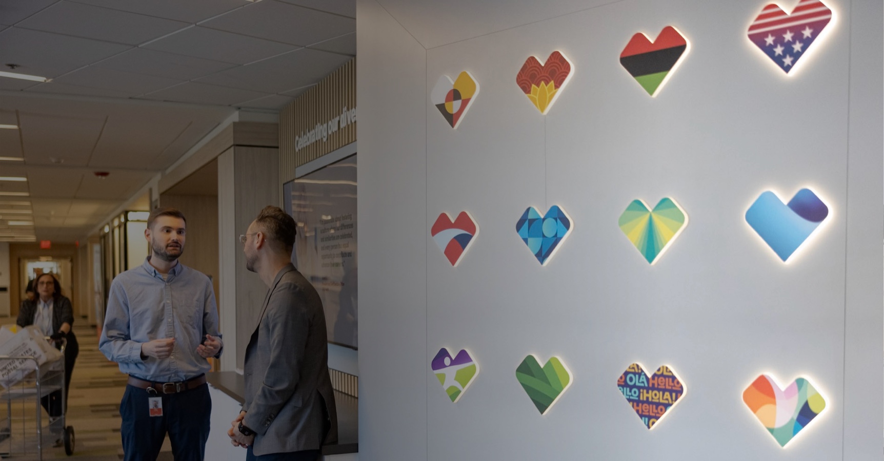 Two men are standing and talking in a hallway next to a wall display with various heart-shaped symbols. Each heart features a different design, including flags and abstract patterns. A woman pushes a cart in the background. The surroundings appear to be a modern facility.