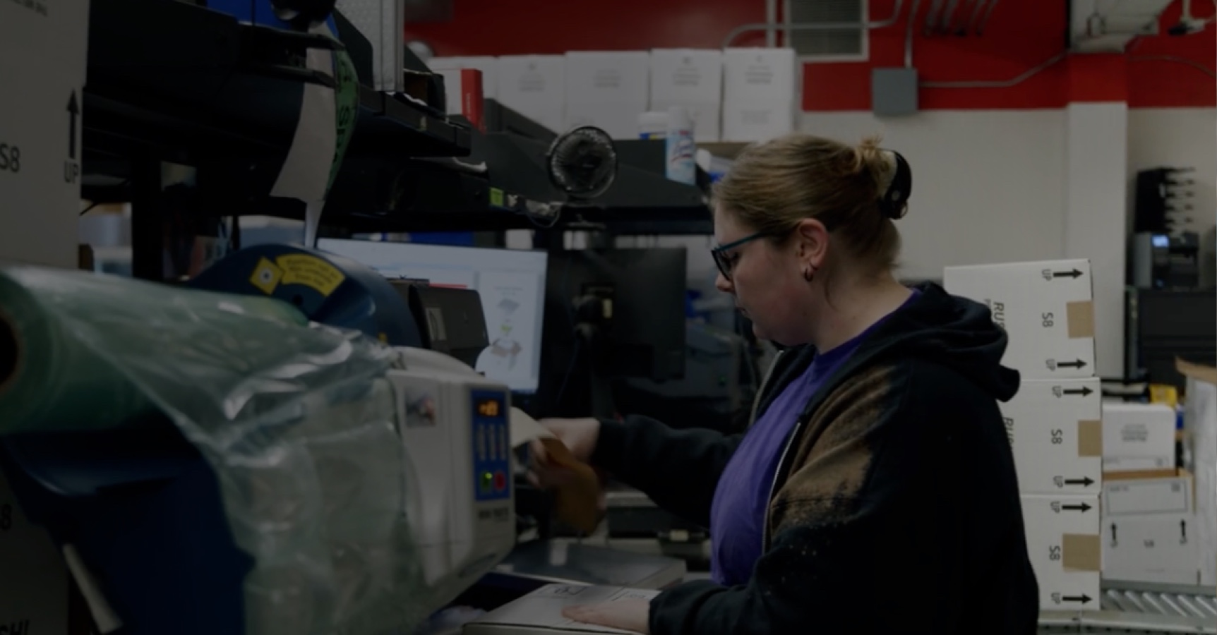 A woman with light brown hair tied back, wearing glasses and a dark hoodie over a purple shirt, works at a machine in a factory or warehouse setting with shelves, boxes, and equipment around her. She appears focused on the task at hand.