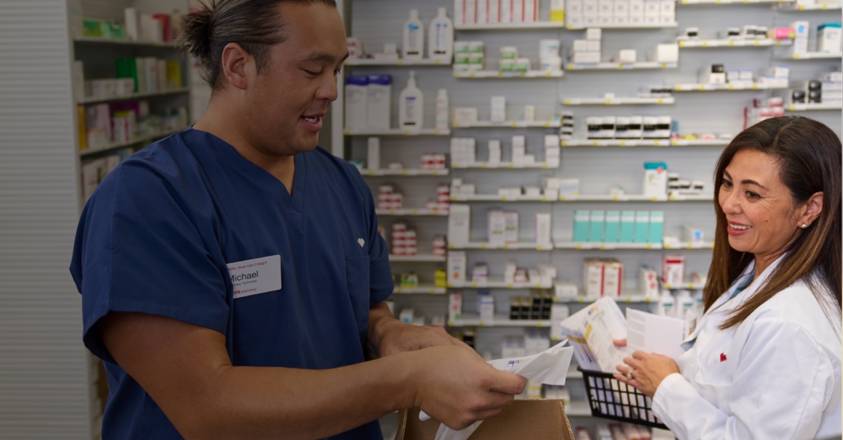 A man in blue scrubs and a woman in a white lab coat work together in a pharmacy. The woman smiles at the man as he opens a box and examines its contents. Shelves filled with medication and products are visible in the background.