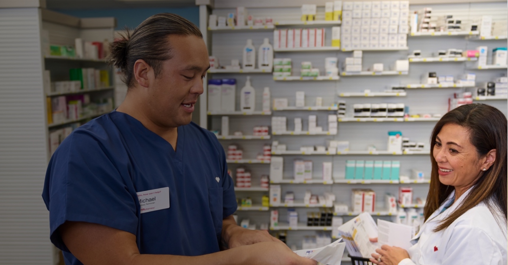 A pharmacist and a pharmacy technician are exchanging documents in a pharmacy. The shelves behind them are stocked with various medications and medical supplies. Both are smiling and appear to be engaged in a friendly conversation.