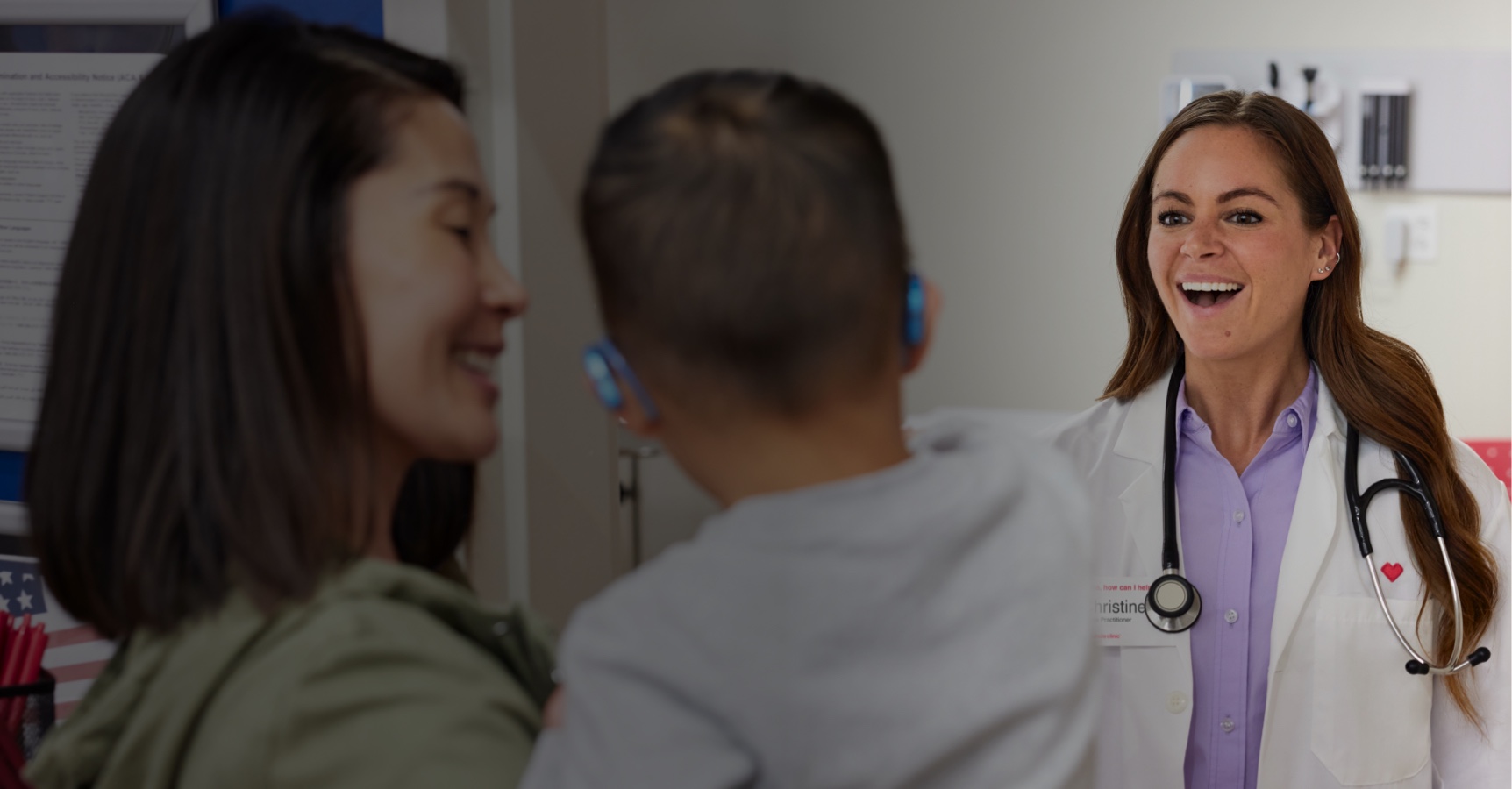 A doctor with a stethoscope smiles warmly at a woman holding a young child with hearing aids in a medical office setting. The woman appears to be talking to the doctor, while the child faces away from the camera. Medical posters are visible in the background.