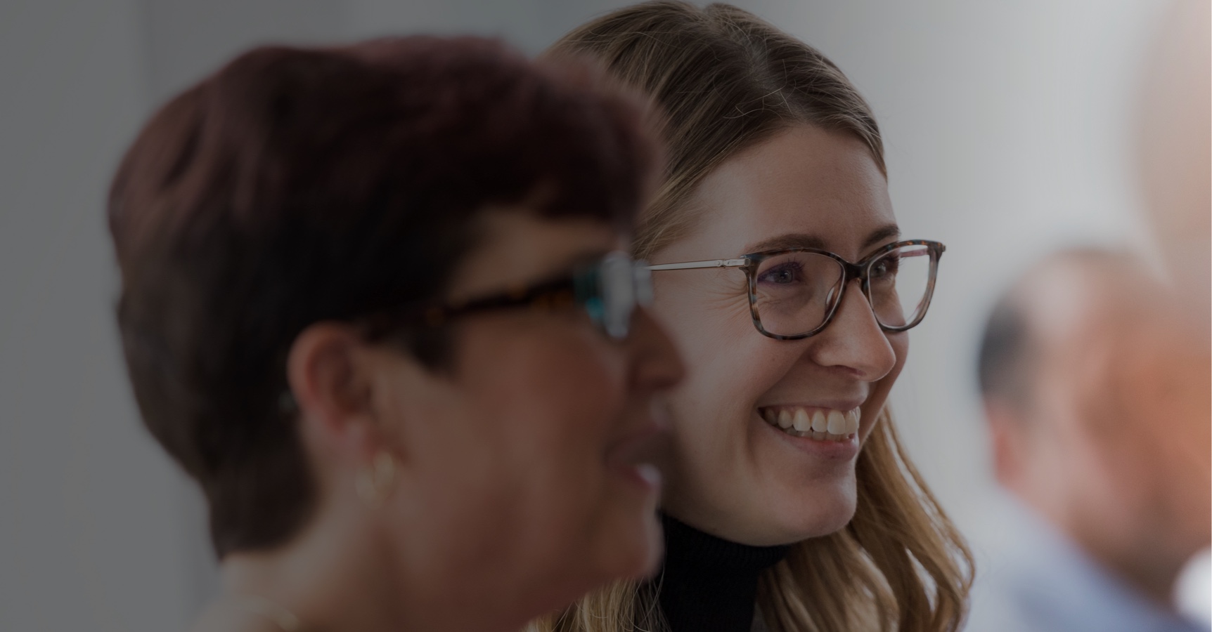 Two people with glasses and smiles are in focus. One has short dark hair, and the other has long light hair. They appear to be engaged in a conversation or event, with a slightly blurred background suggesting an indoor setting.