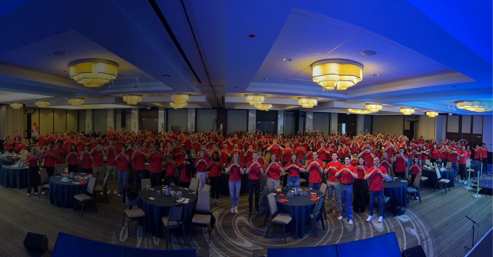 A large group of people, all wearing red shirts, pose together in a conference room. Many are making heart shapes with their hands. There's a backdrop with large, metallic balloons spelling out "IDP." Tables with papers and glasses are visible in the foreground.