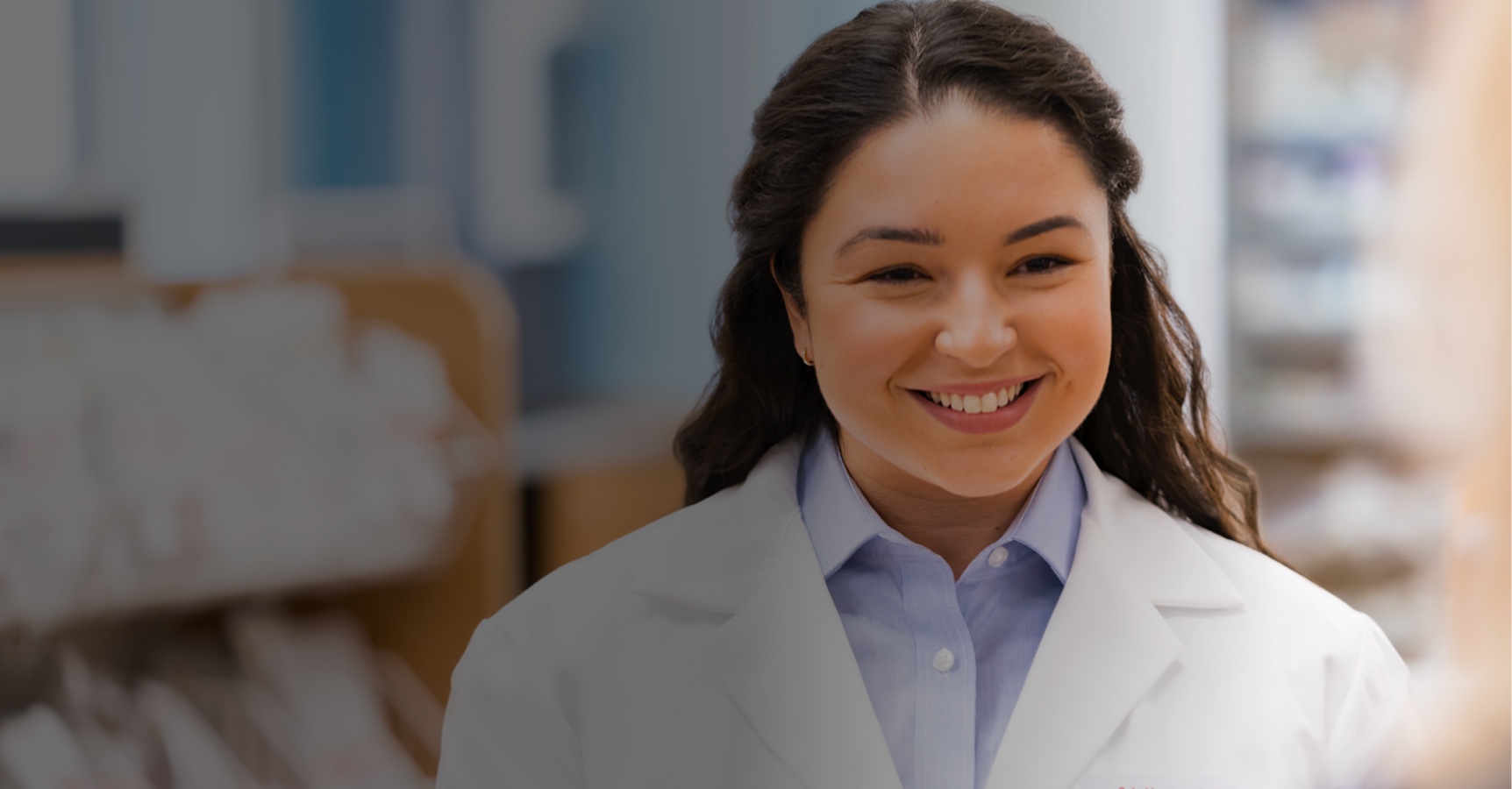 A woman with long dark hair smiles warmly while wearing a white lab coat over a light blue shirt, suggesting she might be a healthcare professional. The background is blurred, focusing on her friendly expression.