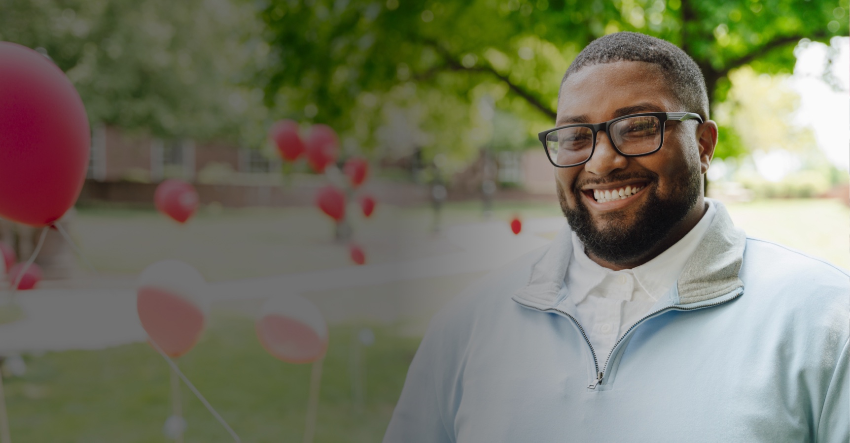 A cheerful man with glasses and a beard is smiling while standing outdoors in a park-like setting. He is wearing a light blue jacket with a white shirt underneath. Red and white balloons are scattered in the background, along with green trees and grass.