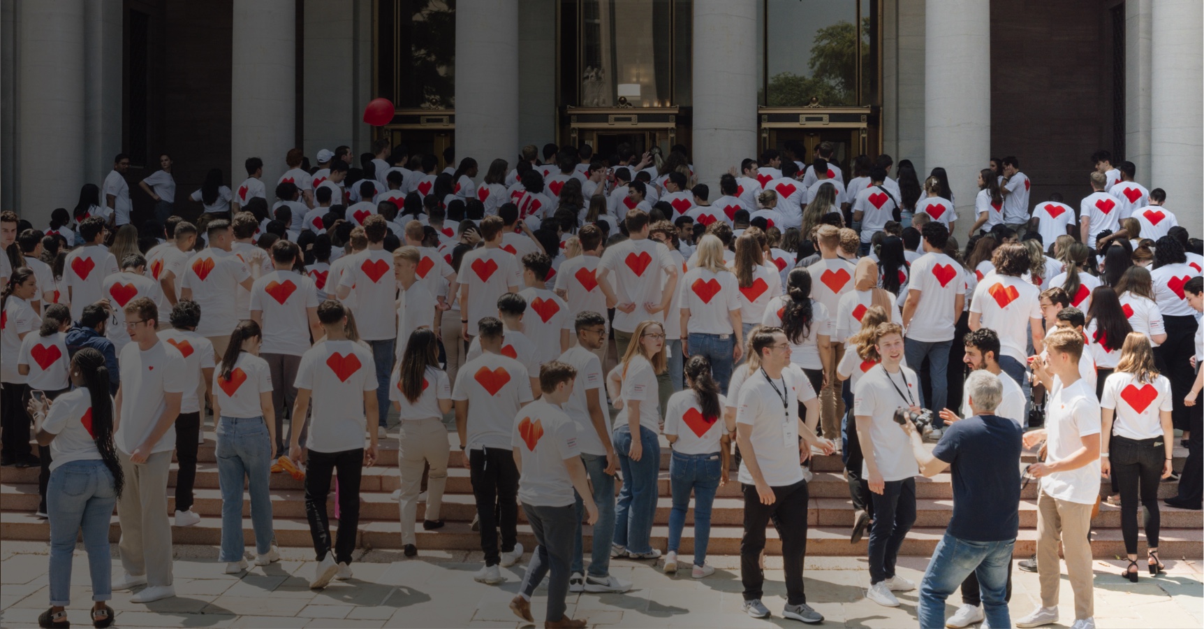 A large group of people wearing white T-shirts with red heart prints on the back is gathered on the steps outside a building with tall columns. Many are standing, socializing, and milling around, while a few are ascending the steps towards the entrance.