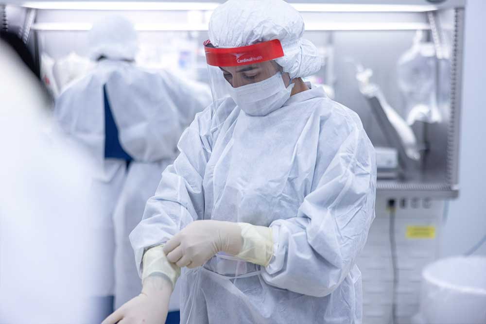 A healthcare professional wearing blue scrubs and a face mask sits beside an elderly woman, also in a face mask. The healthcare worker is showing the elderly woman something on a digital tablet. They are in a bright, cozy room.