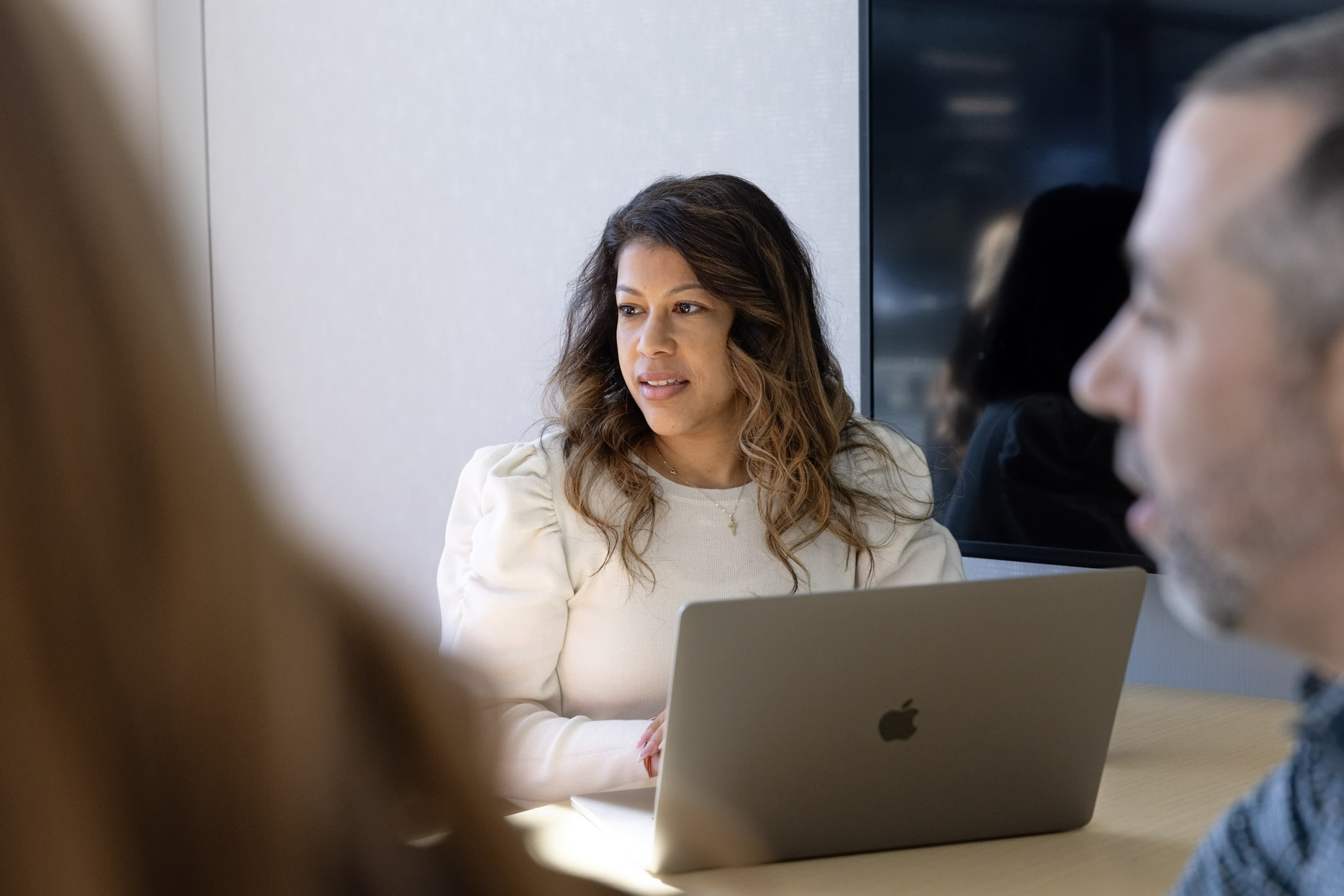 A woman with wavy hair, wearing a white blouse, sits at a table using a MacBook laptop. She looks attentively towards the left. A blurred man is in the foreground on the right side, and a blurred person is partially visible on the left.